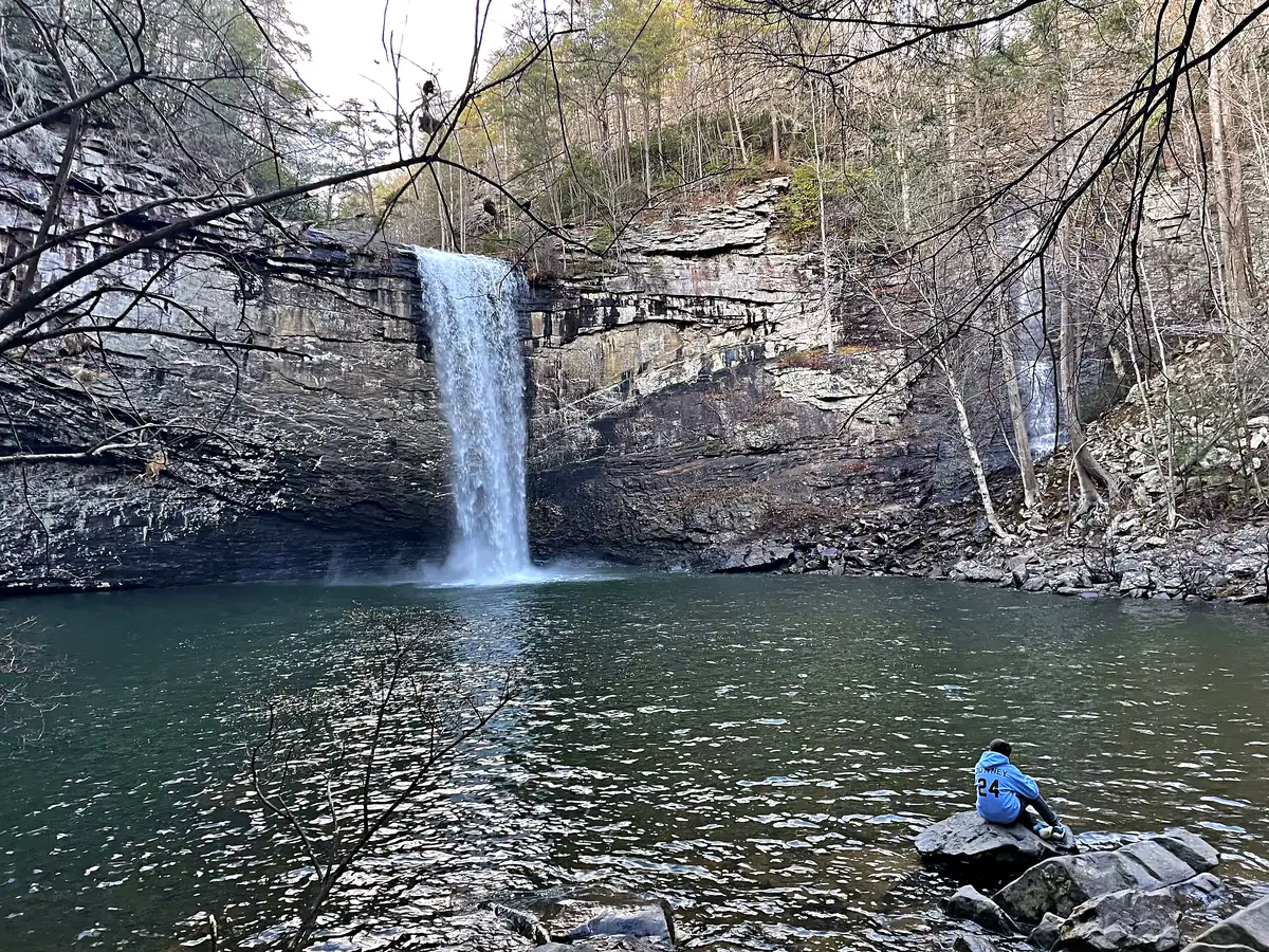 Foster Falls, a 60-foot waterfall pouring off a layered rock cliff into a wide green pool, with one of our kids sitting on a boulder at the edge of the water